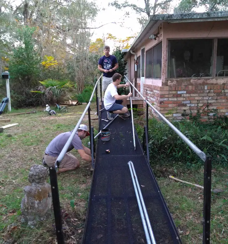 Three volunteers are almost finished constructing a black metal ramp on an old brick house.